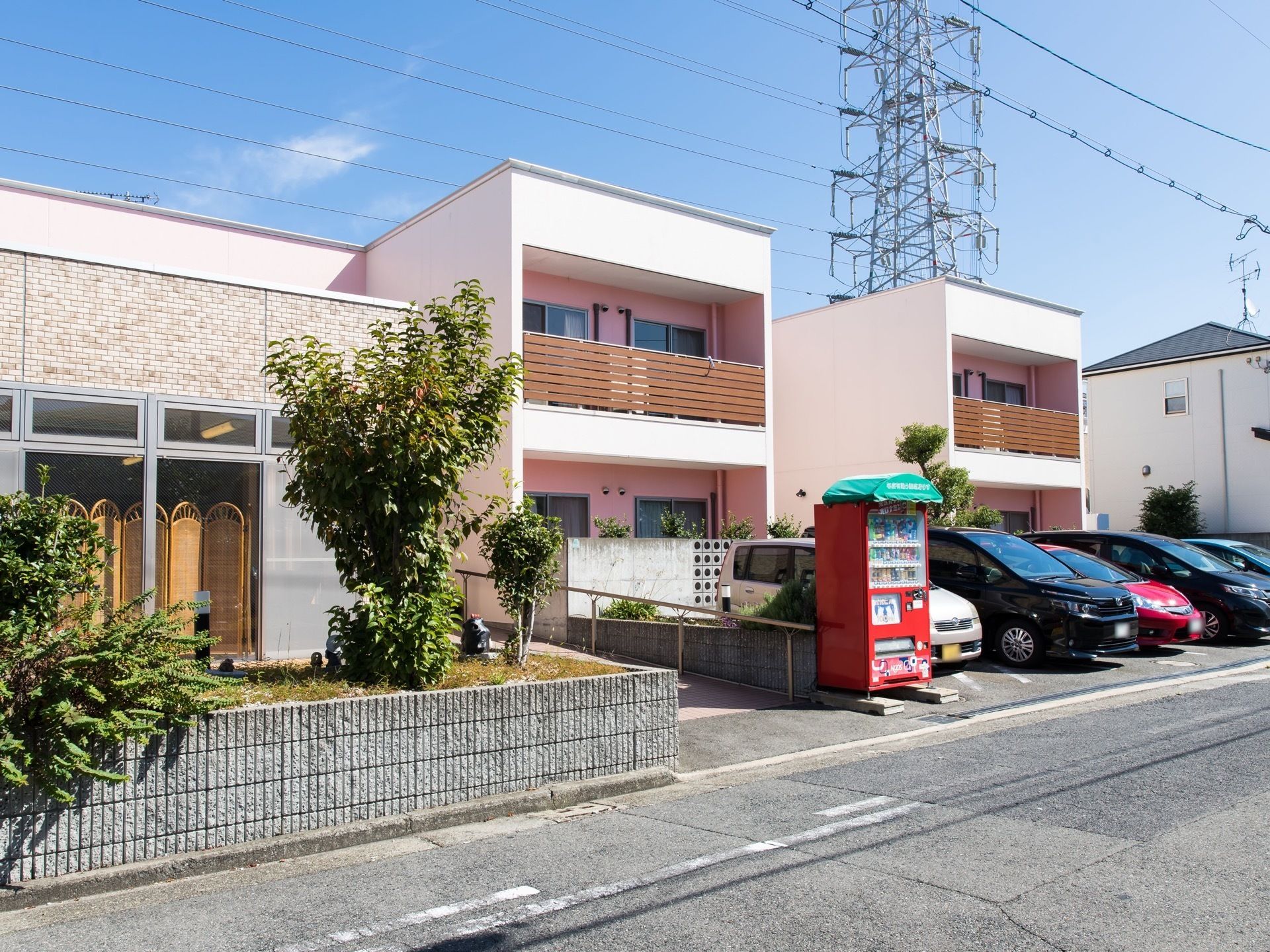 ピンク色の壁面の２階建ての建物がある。建物の前には駐車場がある。玄関へと続く道には手すりがある。