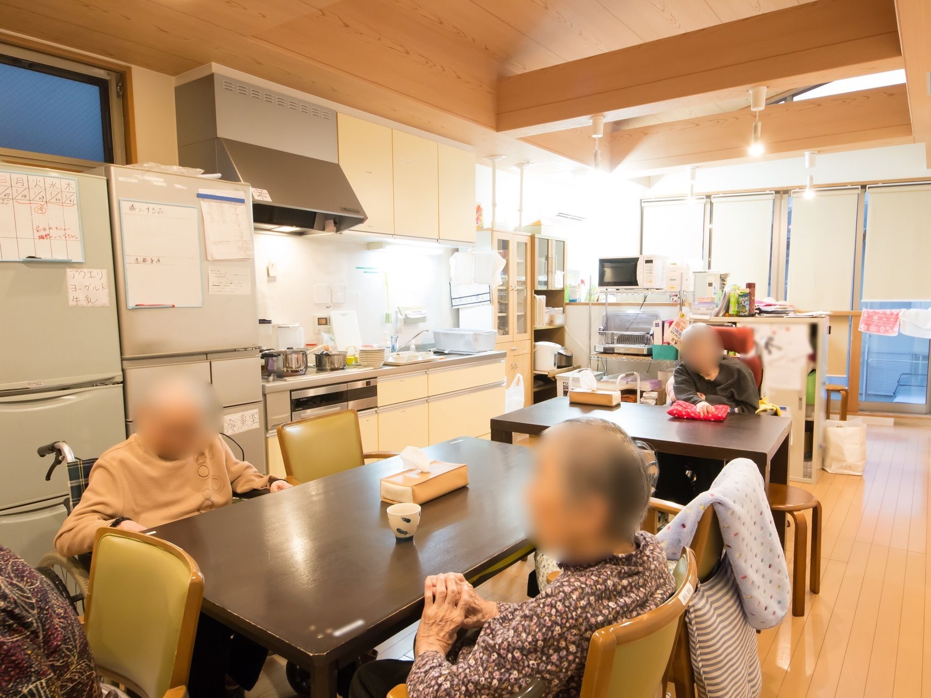 食堂にはキッチンスペース、2台の冷蔵庫がある。同じスペースにテーブルと椅子が並べられている。席には入居者様が座っている。