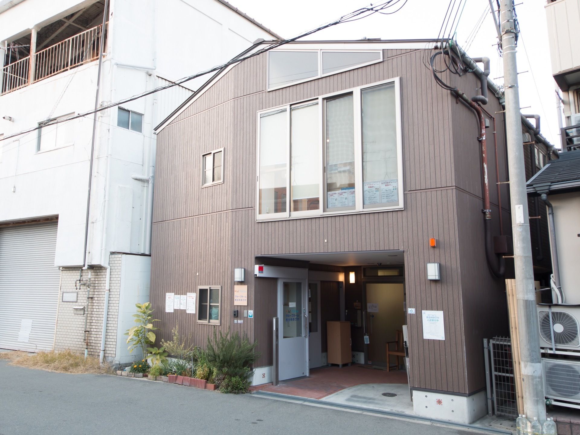施設は茶色で一軒家のような外観で、奥行きのある造りである。玄関前には複数のプランターが並べられている。
