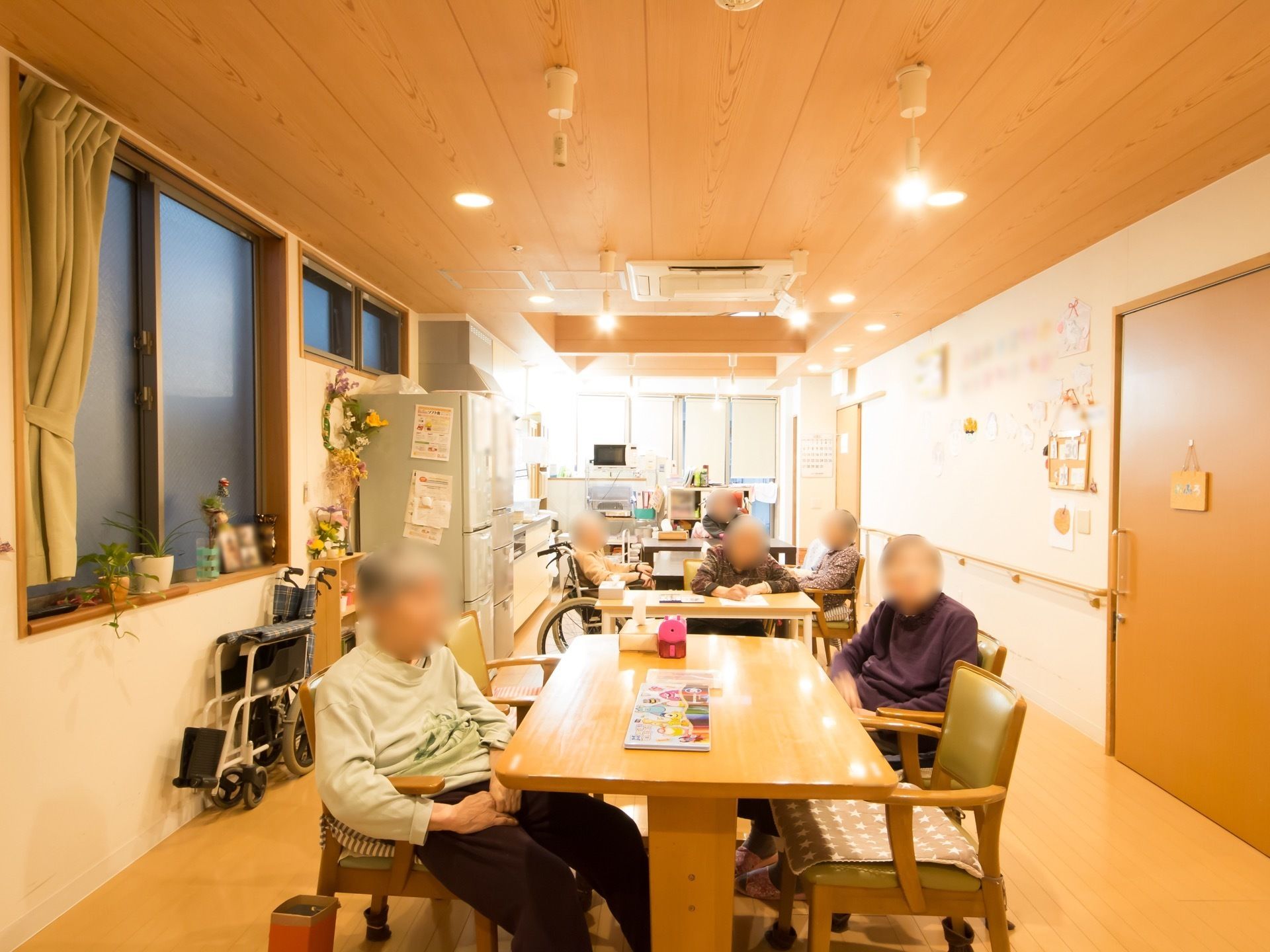 電球色の照明が温かく、木のぬくもりが感じられる食堂。２台の大きなテーブルが置かれており、奥には冷蔵庫や調理台が設置されている。