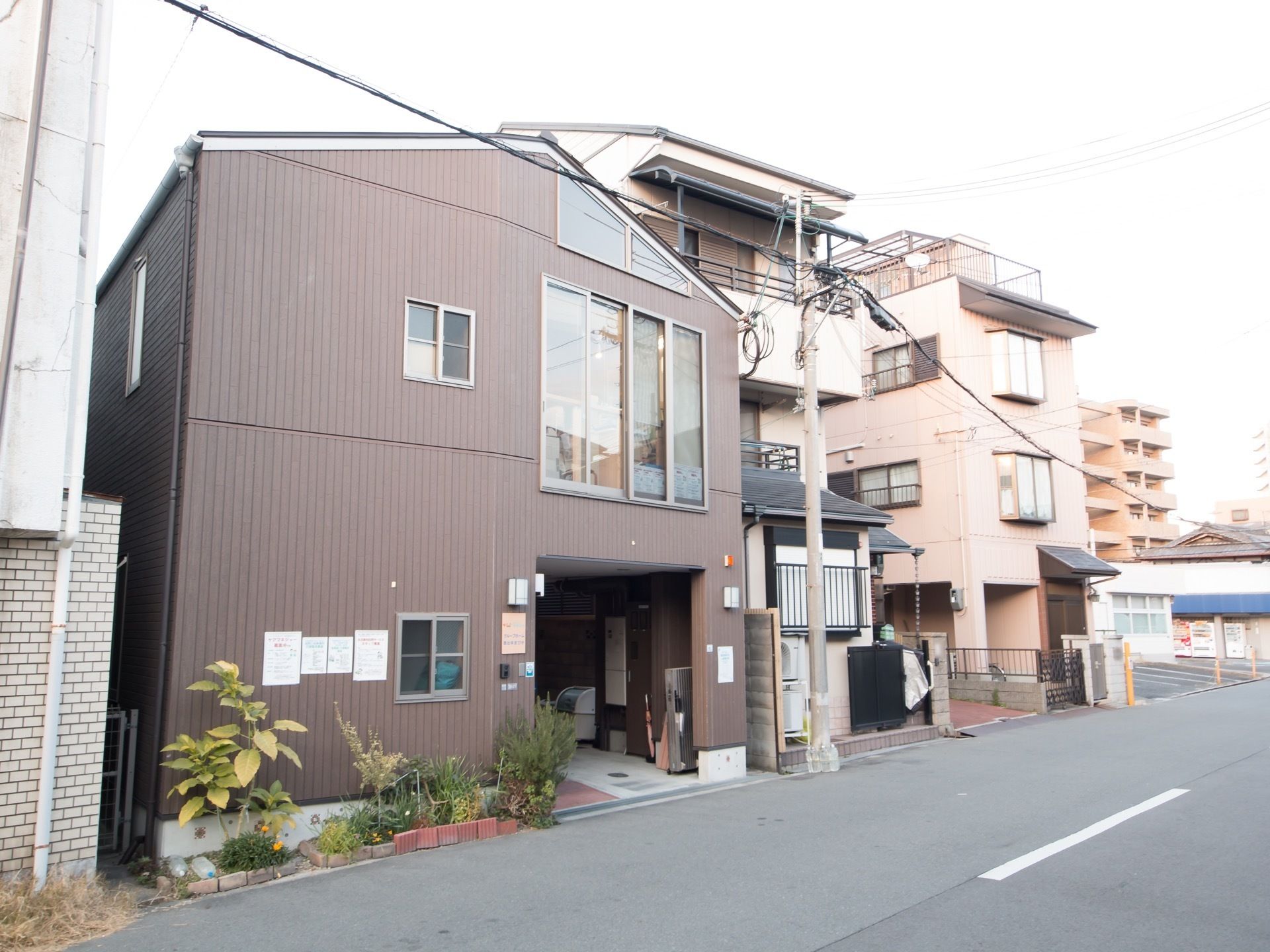 どっしりとした印象を受ける焦げ茶色の外壁をした、木造造り２階建ての建物。道路に面した壁際に花壇が作られている。