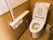フローリングに木材を使用した壁の個室トイレである。中央に温水洗浄便座が設置された洋式トイレがあり、壁に非常ボタンがある。