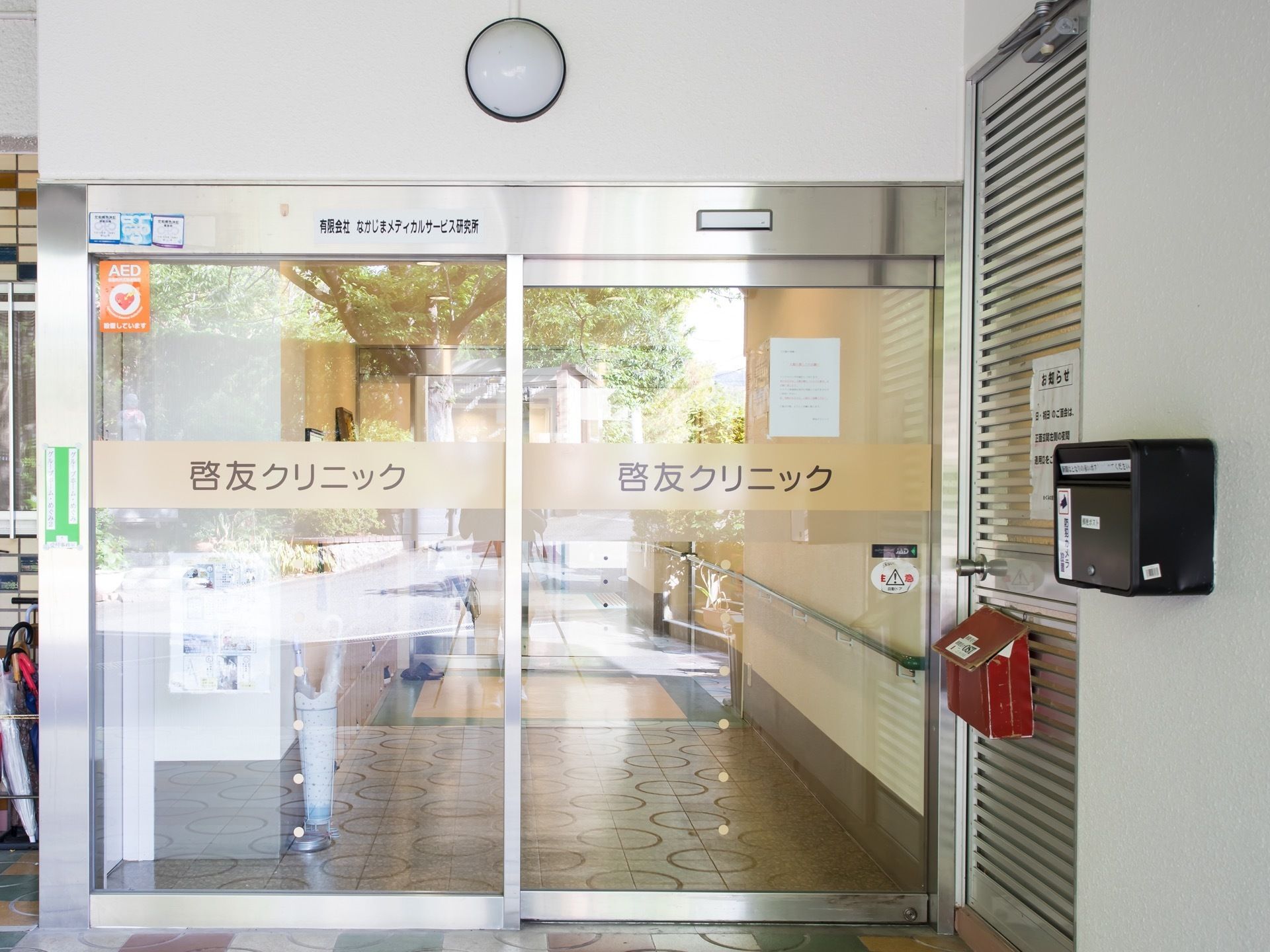 ガラス製の自動ドアがあり、施設名が書かれている。ドアの横の壁には赤と黒の郵便受けが１つずつ取りつけられている。