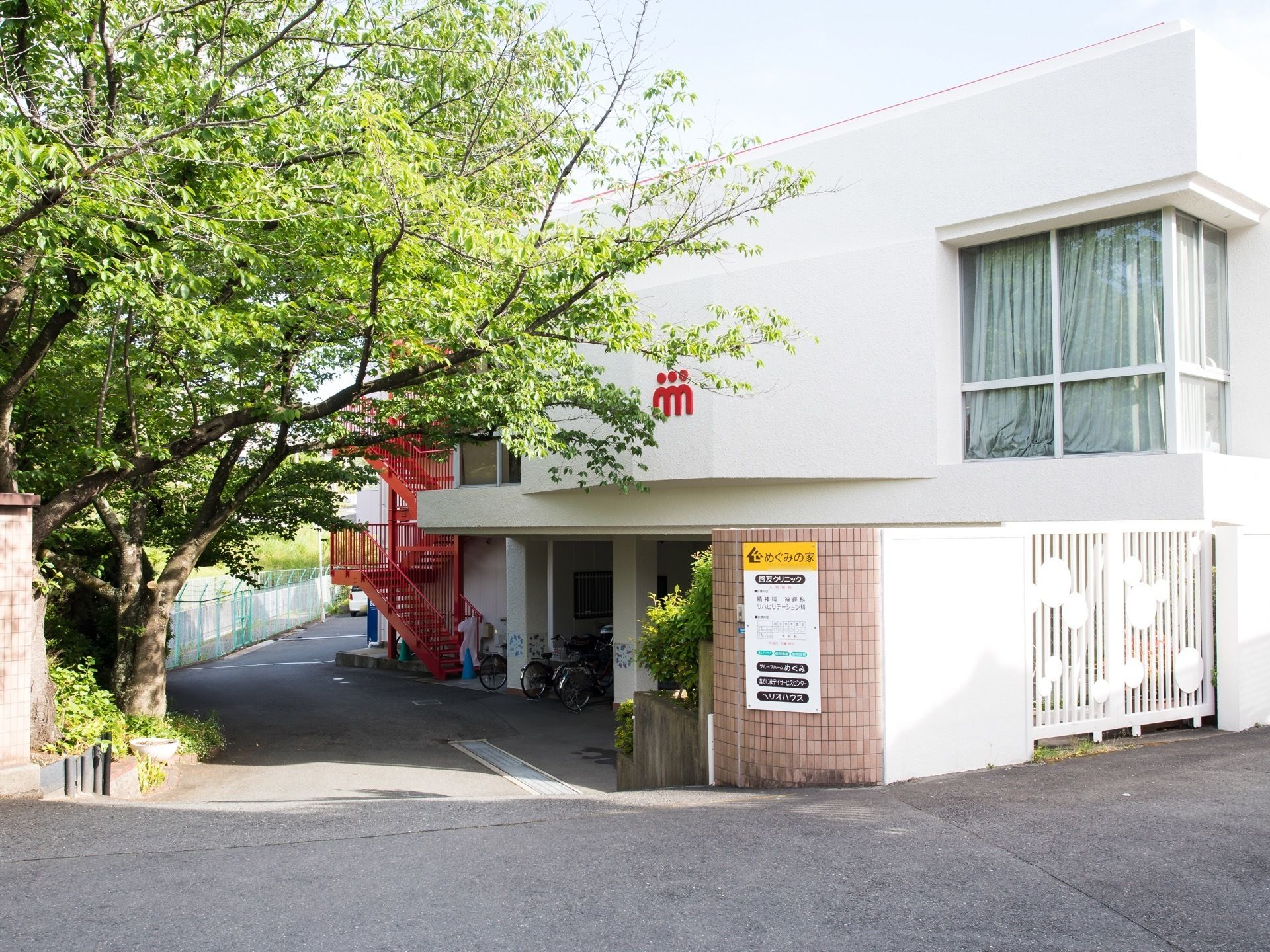 白い建物の前には、複数の施設名の書かれた看板が取りつけられたブロック塀がある。また、奥には赤色の外階段が見える。
