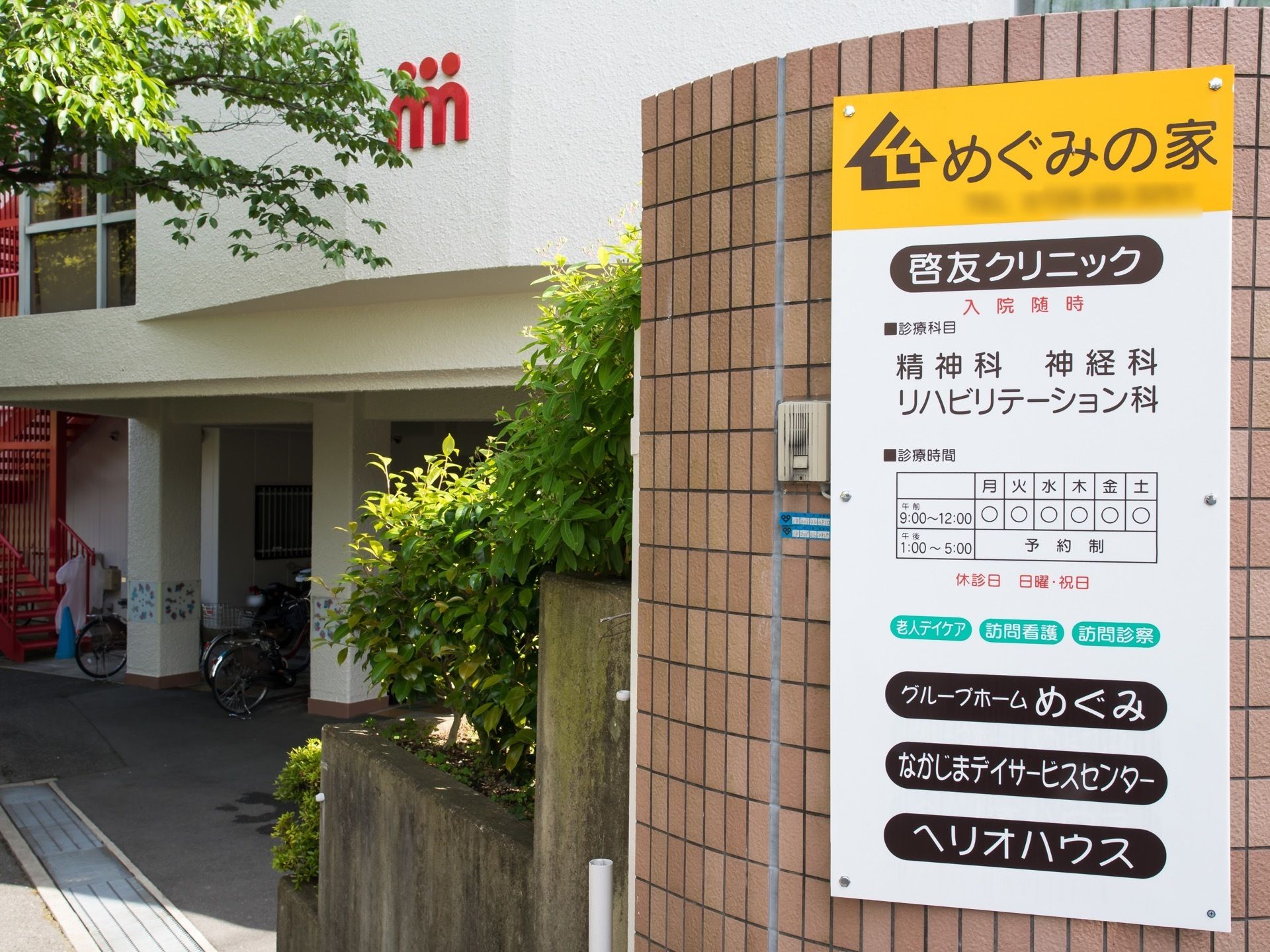 複数の施設名が書かれた看板がある。茶色のブロック塀に貼られており、その横にインターホンが設置されている。