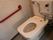 蓋のない洋式トイレである。便器には黒色で注意事項がかかれており、壁には赤色の手すりが取りつけられている。