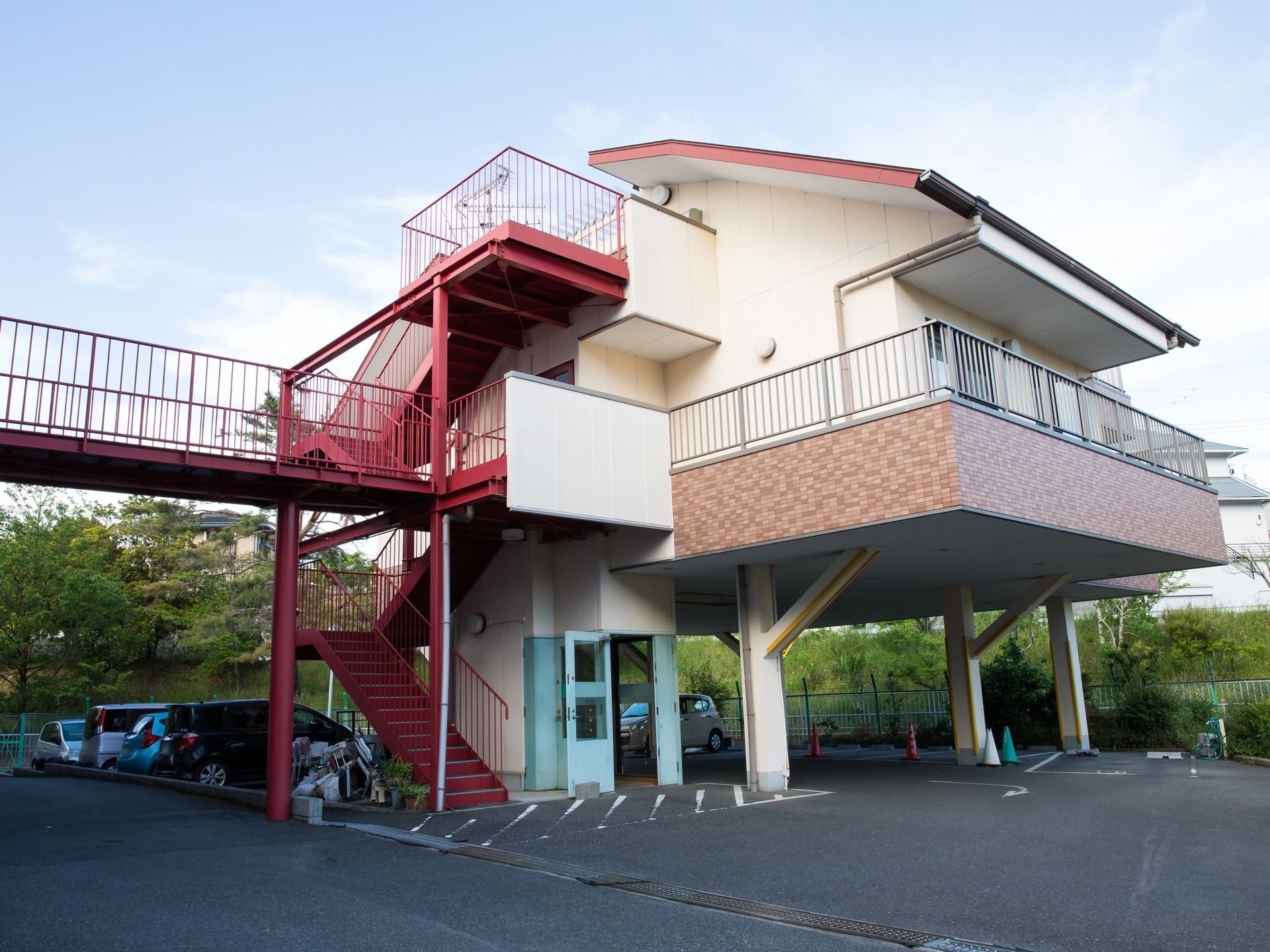 １階部分が駐車場となっている建物である。駐車場には車が複数停まっており、赤色の外階段と通路が設置されている。