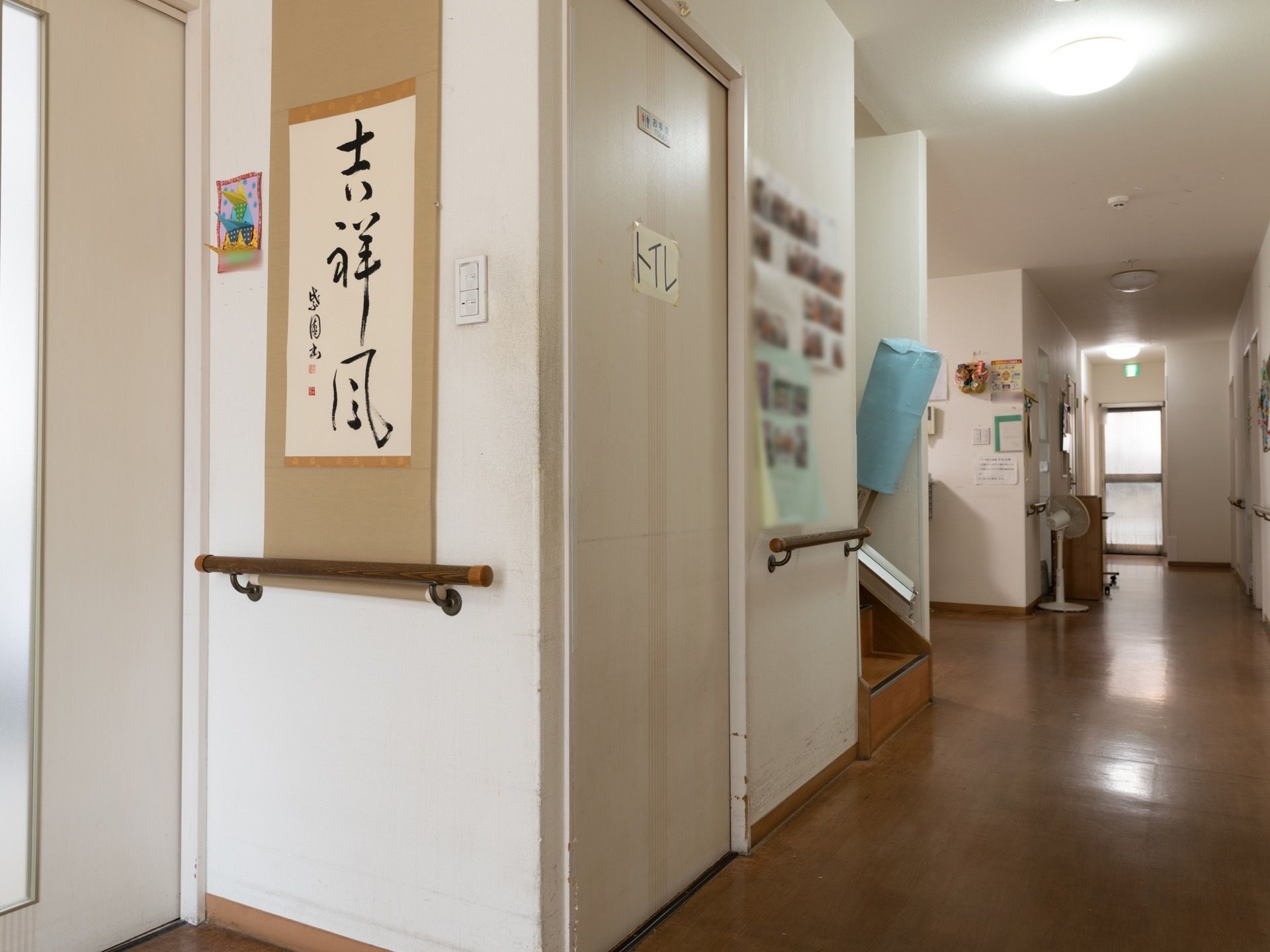 階段のそばにはトイレがある。トイレのドアにはトイレと書かれた紙が貼ってある。近くの壁には入居者様の写真が掲示されている。