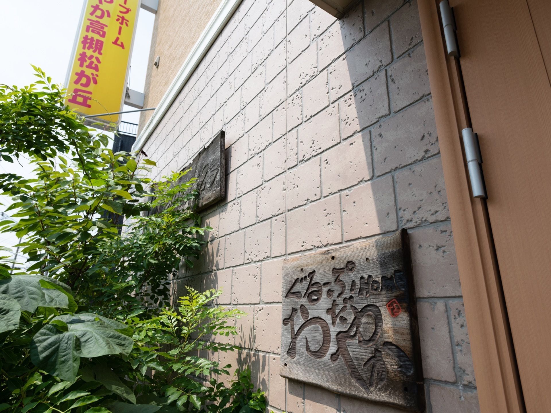 施設の入口そばの壁には、プレートが掲げられている。木の素材でできており、施設の名前がひらがなで記されている。