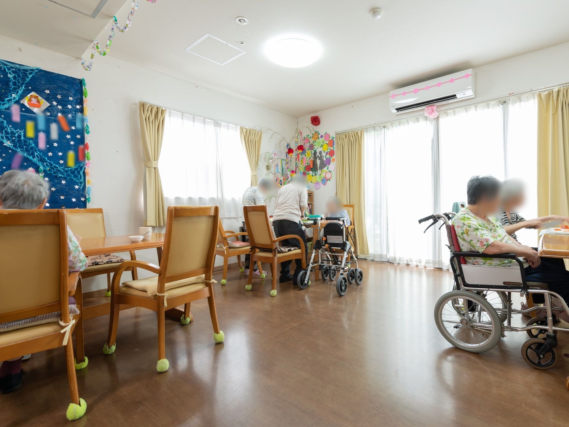 窓が大きいので、日中は光がよく入り明るい。エアコンが設置されている。入居者様が車椅子や椅子に座り、作業をしている。