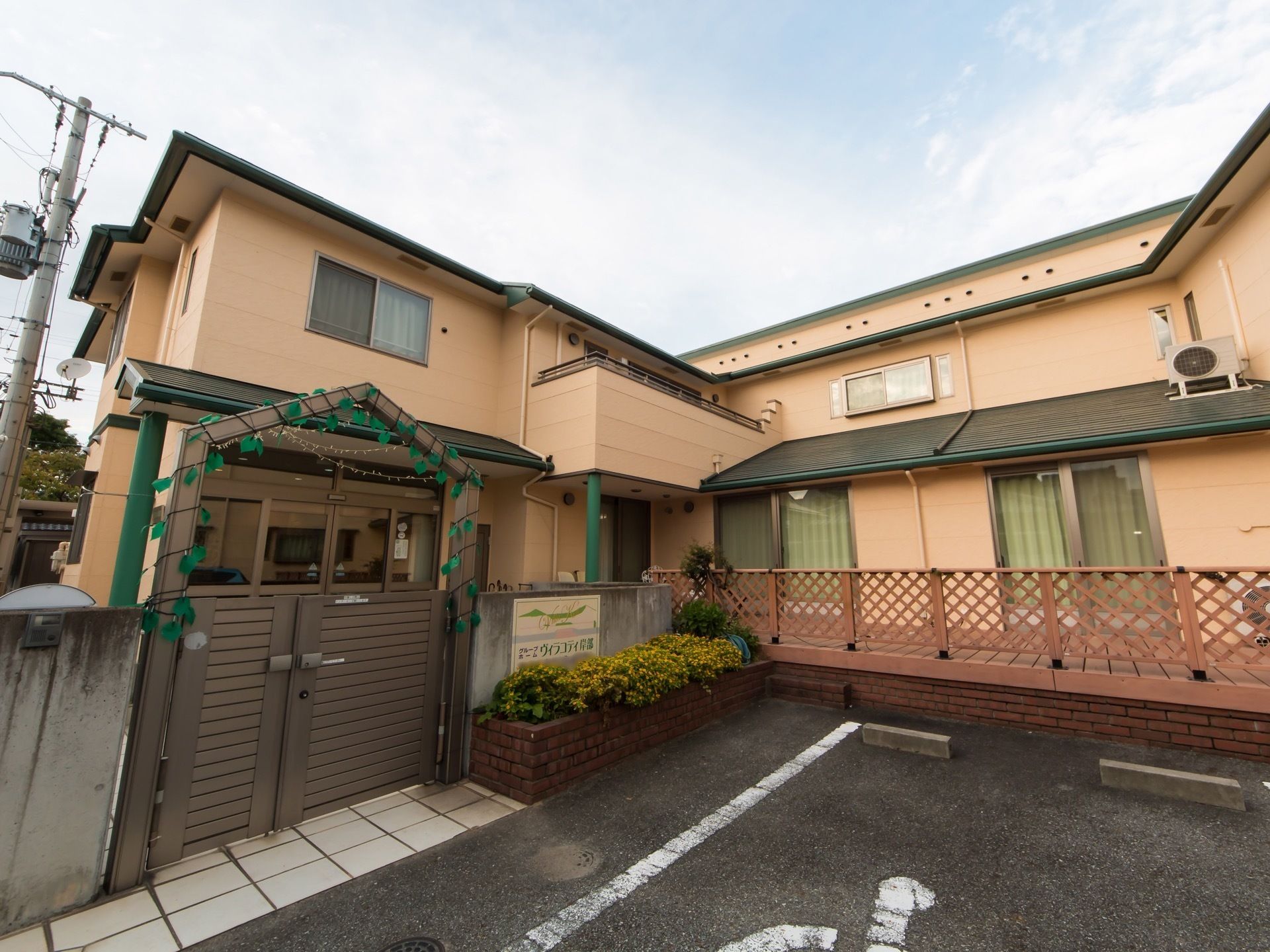 ベージュの壁とダークグリーンの屋根の２階建ての建物がある。コンクリートと木製の塀で覆われ、玄関前には樹木が植えられ、側に駐車場がある。