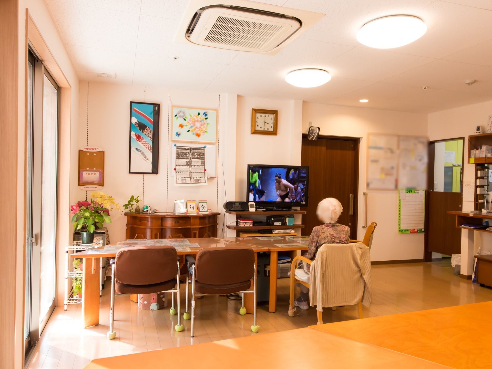 壁側にテレビが置かれ、テレビに向かって座っている入居者様の様子である。周囲の壁には、たくさんの掲示物や飾り物がある。