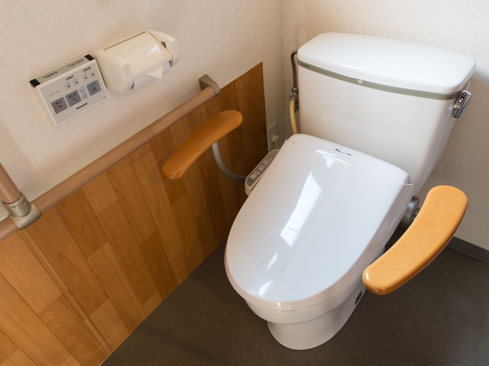 タンクつきの便器が中央に置かれ、手すりが壁と便器の両側に取りつけられている。また、壁にはトイレットペーパーとリモコンがある。