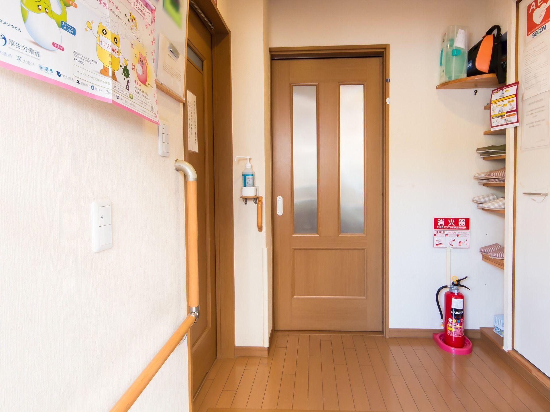 扉の前の床には消火器が置かれており、壁には消毒液が備えつけられている。また、スリッパが収納されている棚もある。