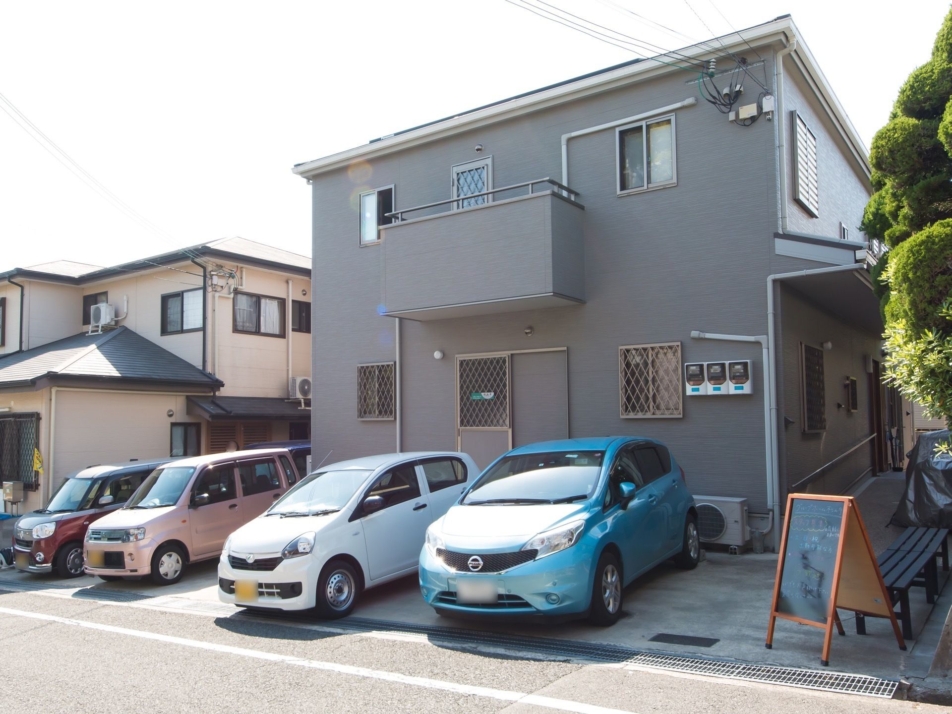 グレーの壁の２階建ての建物である。２階の一部にはベランダがあり、１階の窓には格子がつけられている。また、車が数台駐車してある。