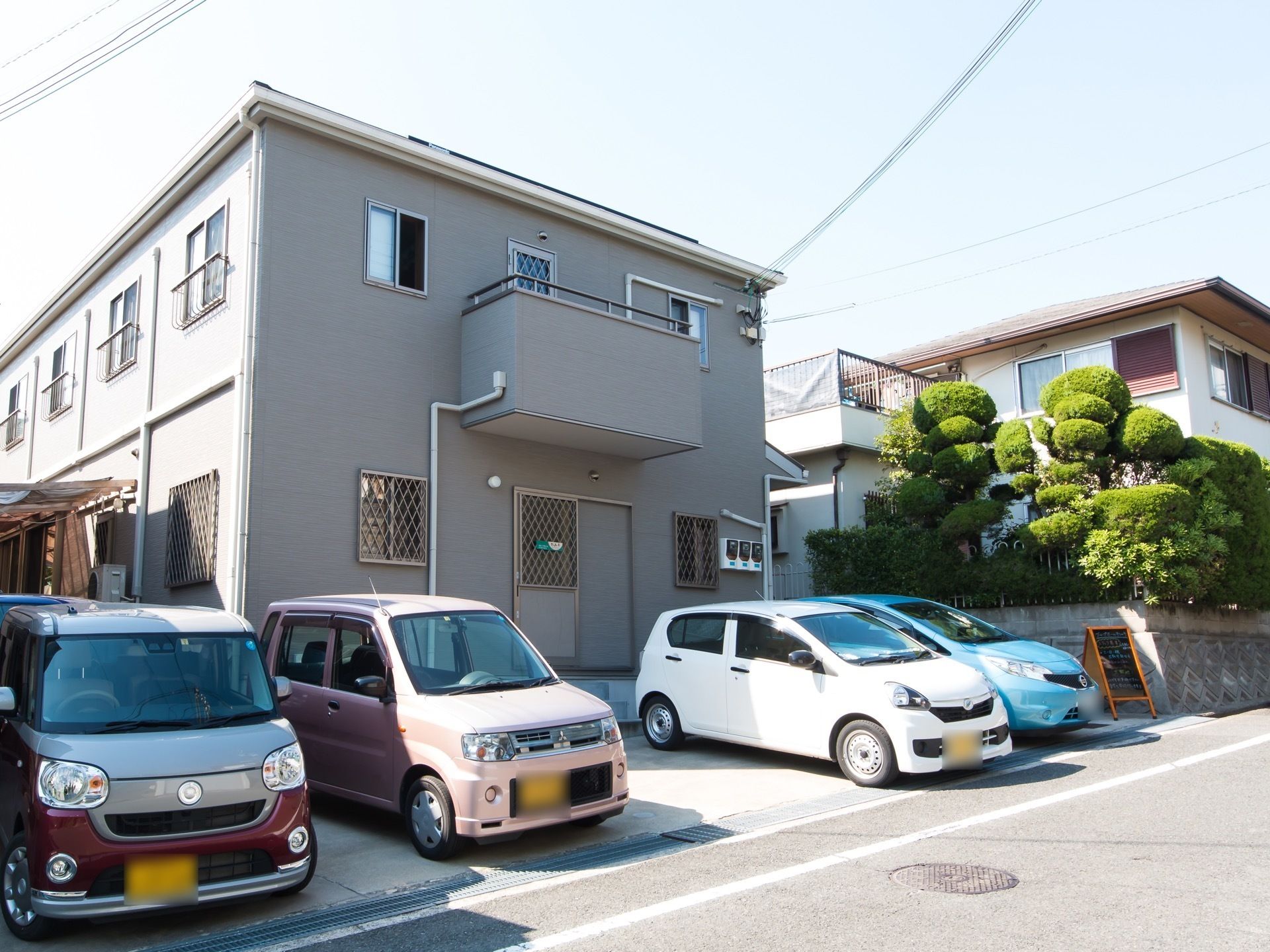 ２階建てのグレーの建物である。２階の窓には手すりが設置されており、一部ベランダもある。車が数台駐車している。