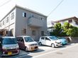 ２階建てのグレーの建物である。２階の窓には手すりが設置されており、一部ベランダもある。車が数台駐車している。