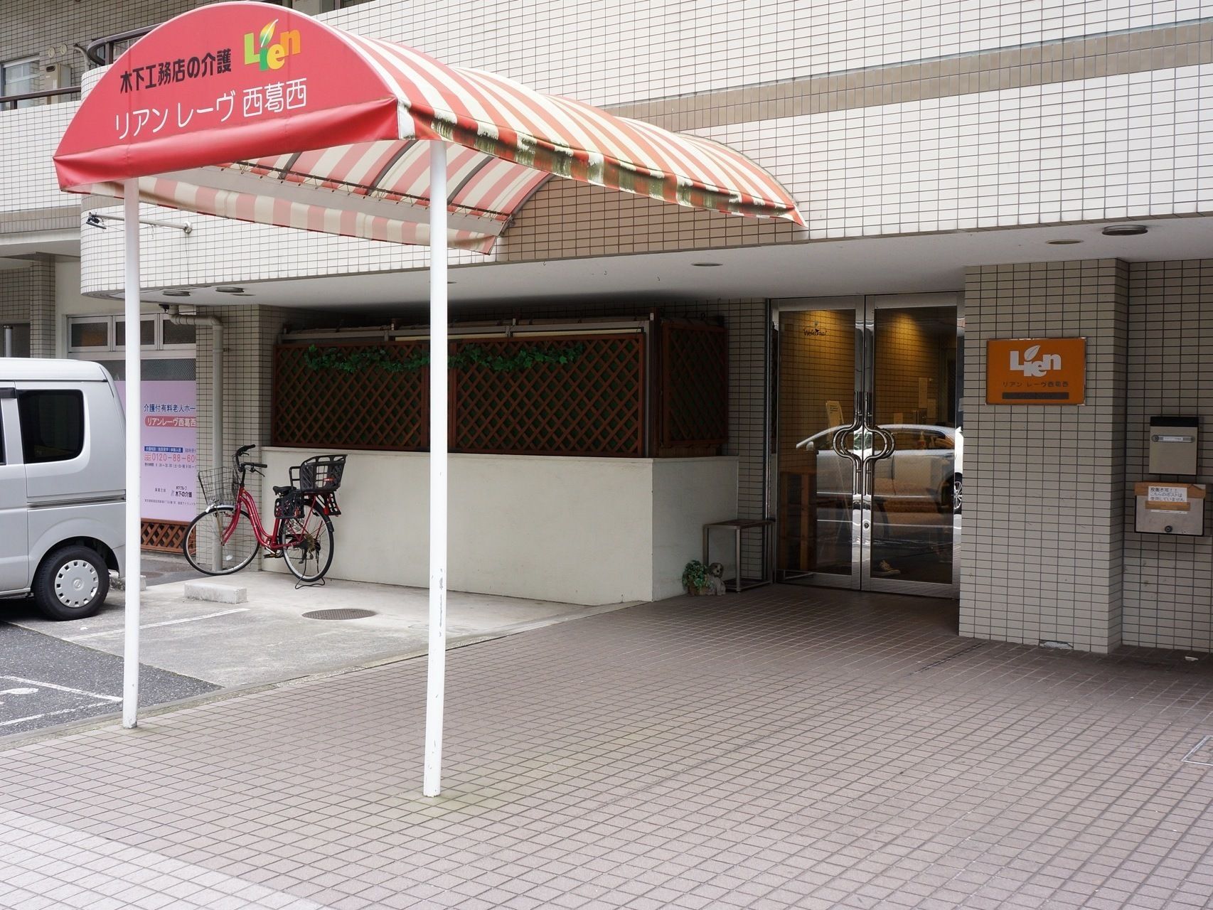 前面に施設名が書かれたストライプ模様の屋根が、駐車場のすぐ横から玄関ドアの前まで設置されている。左端に白い車がある。