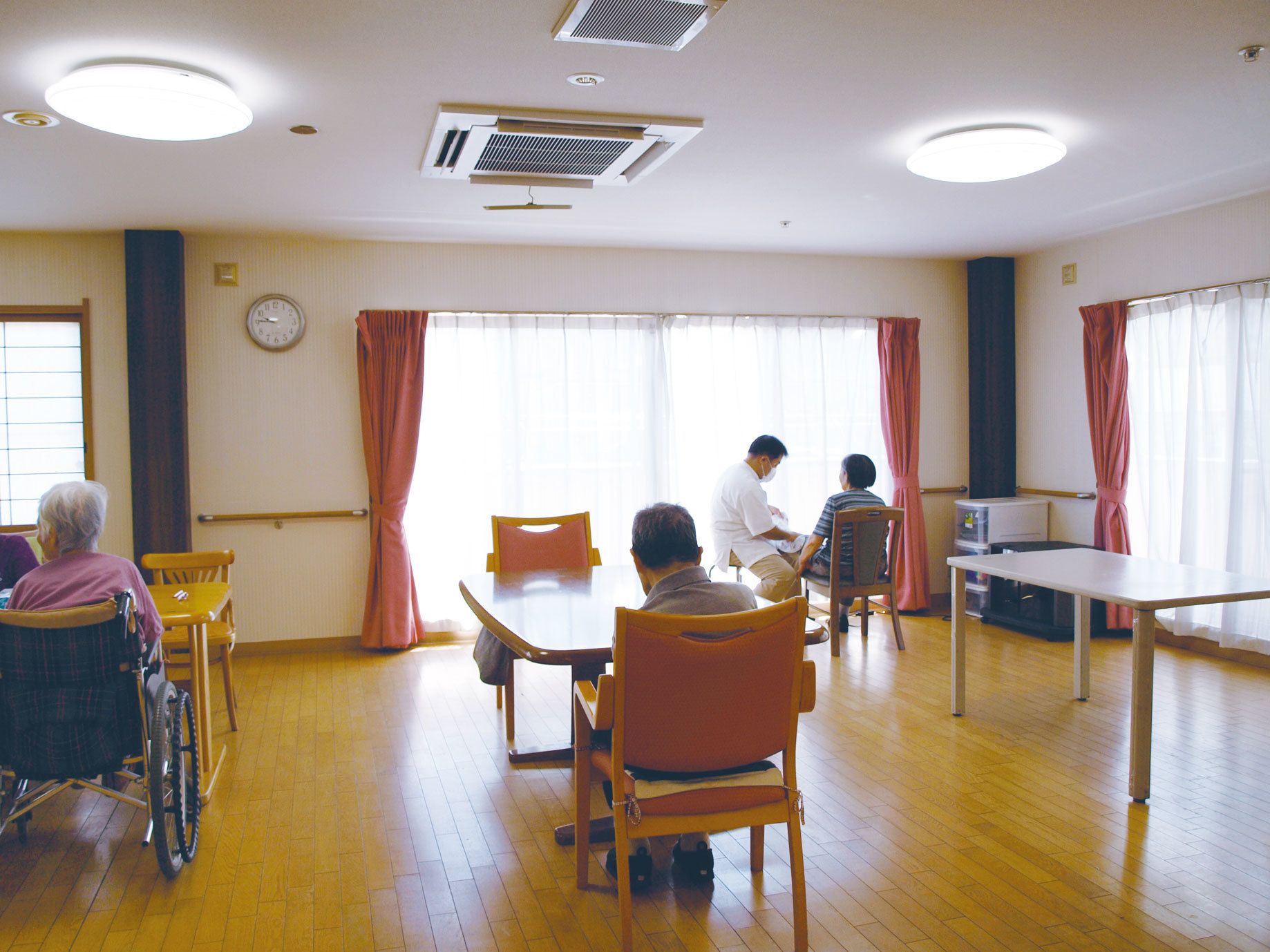 窓が大きく明るい部屋に置かれたテーブルと椅子でくつろぐ入所者たち
