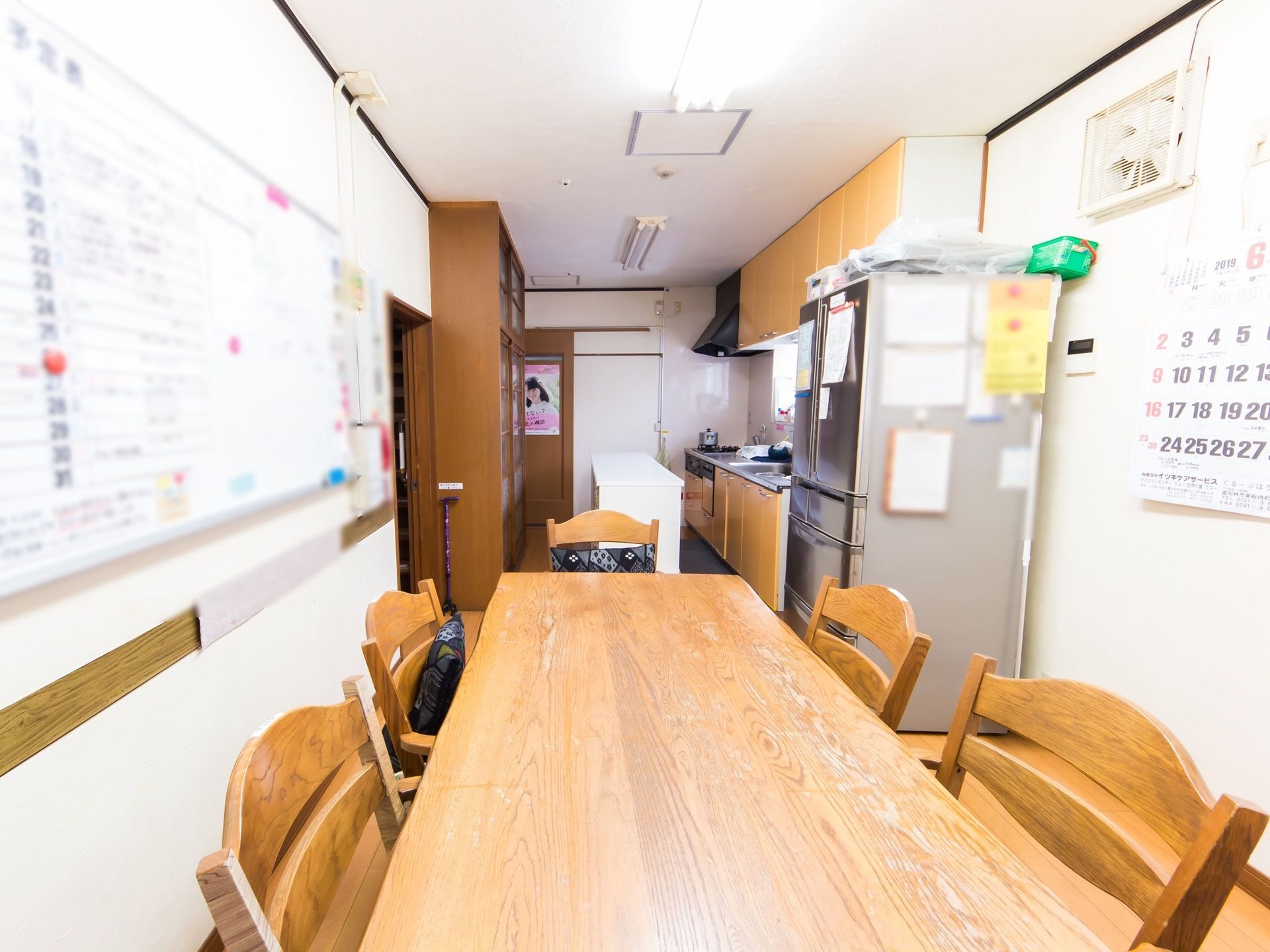 ダイニングの木製テーブルの各面に椅子が並べてある。壁には、カレンダーやホワイトボードのスケジュール表がある。