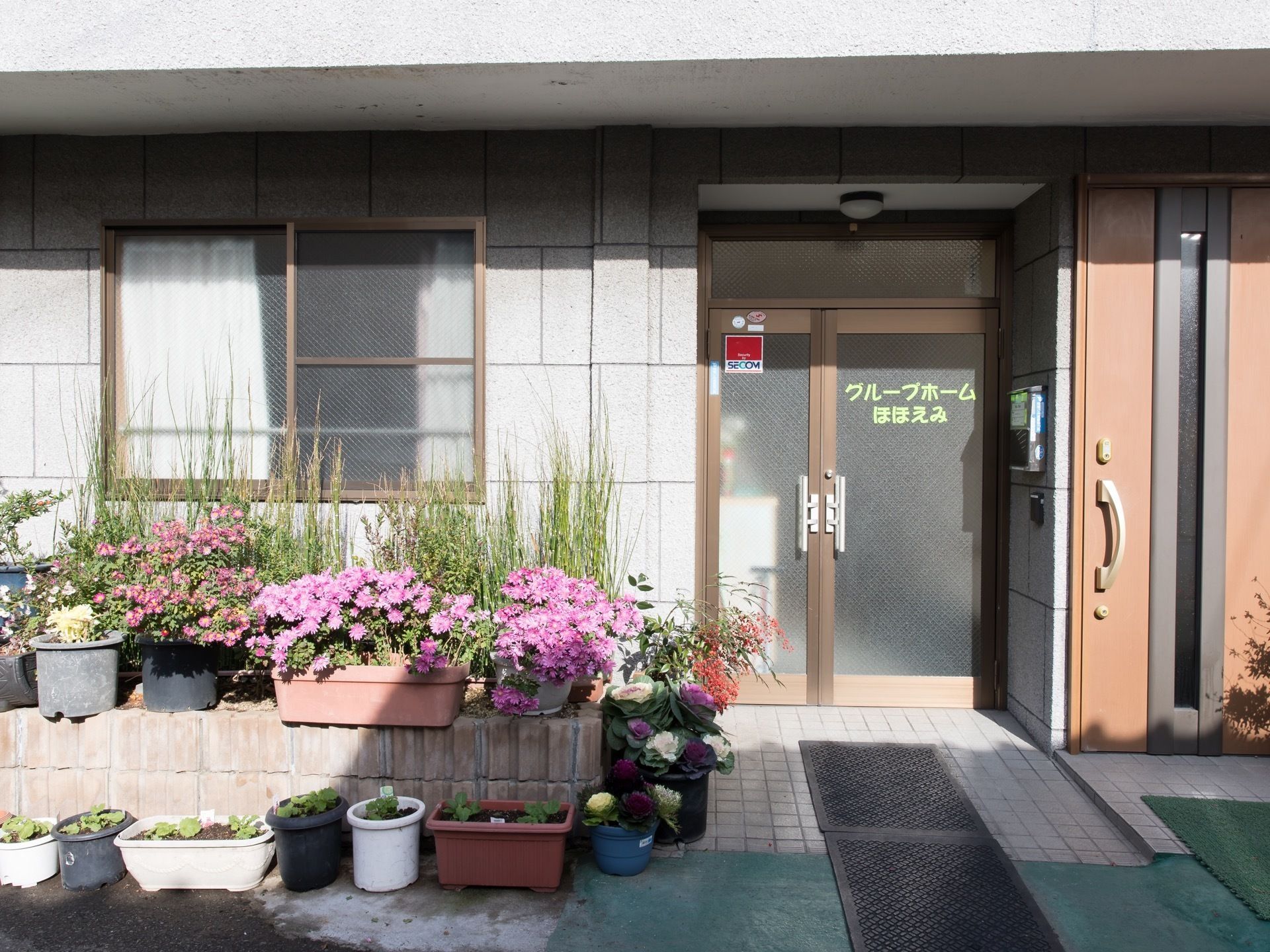 施設入口の図である。正面右側に片開きの玄関扉があり、壁にポストが付けられている玄関前にたくさんの花が飾られている。