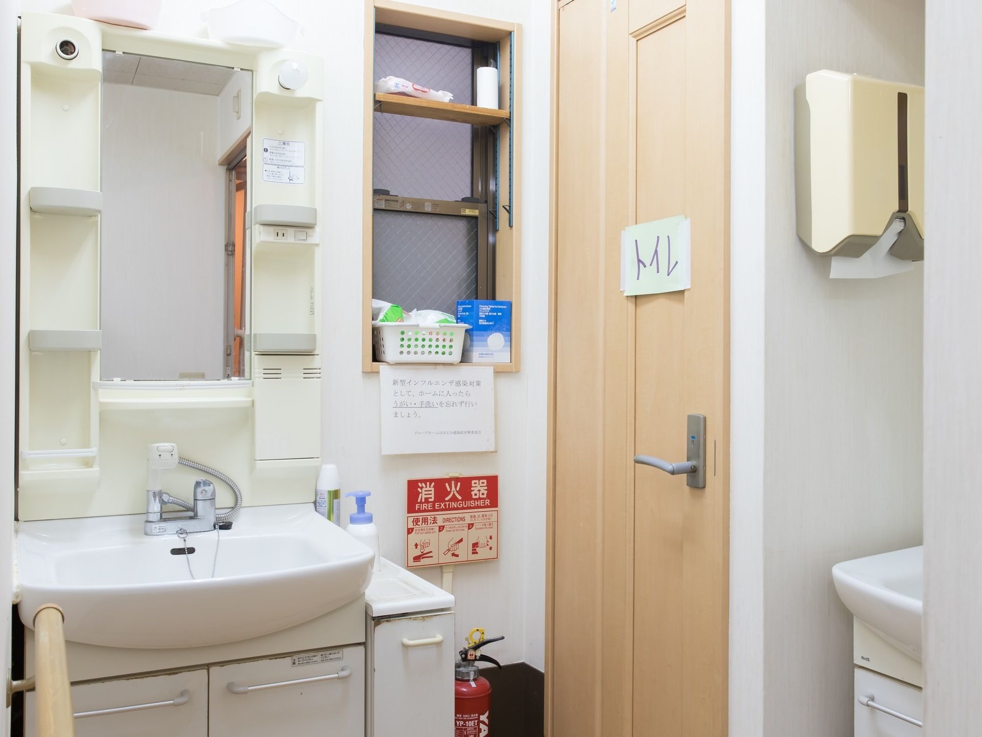 トイレが併設された洗面台である。左正面に白い洗面台があり、壁に鏡が付けられている右手にトイレへのドアがある。