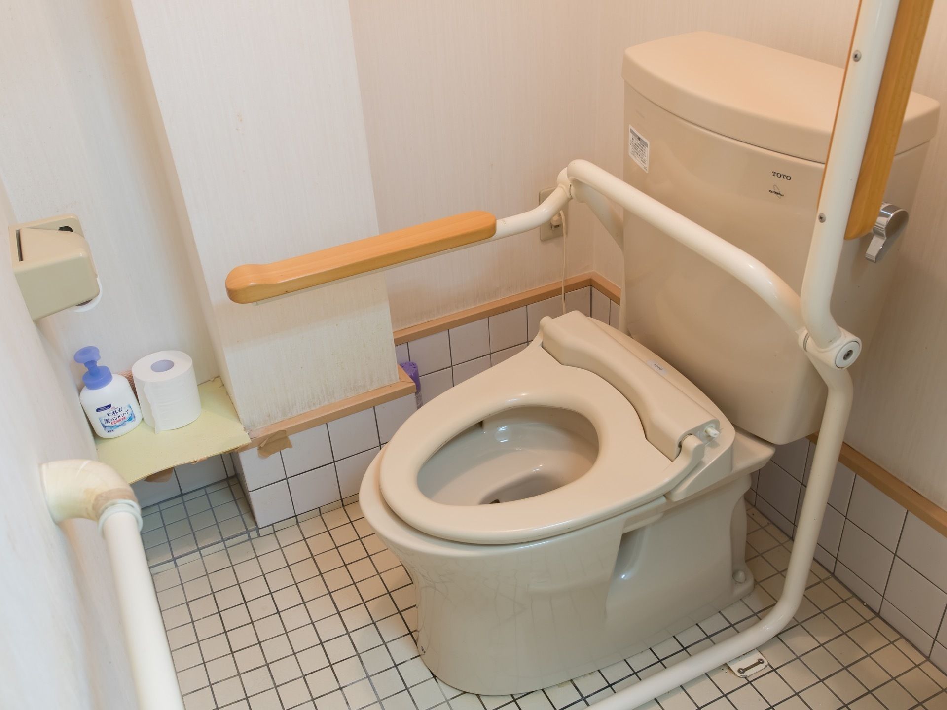 白いタイルが床と壁に貼られた個室トイレである。中央に温水洗浄便座が設置されたトイレがあり、手すりが用意されている。