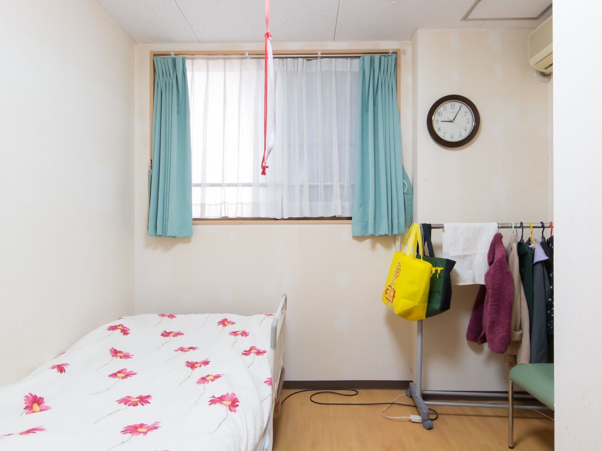 白い壁の居室で、左側の壁際に布団が敷かれたベッドがある。正面に青いカーテンが掛けられた窓がある。