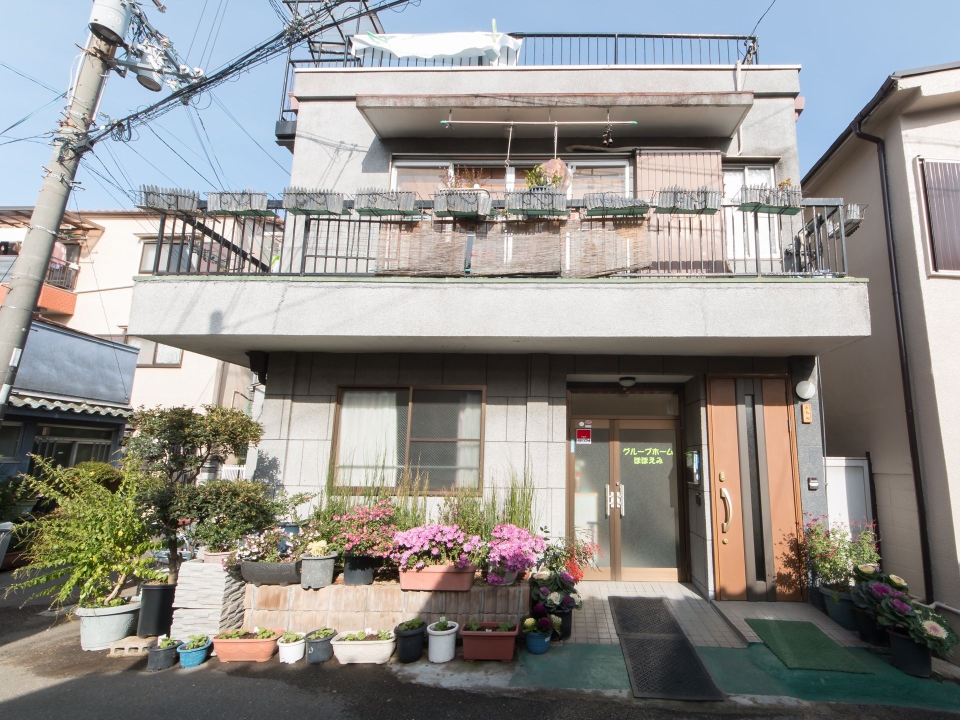 施設は２階建てで、２階部分にバルコニーがあり、その上には屋上も設けられている。入口前にたくさんの花が飾られている。