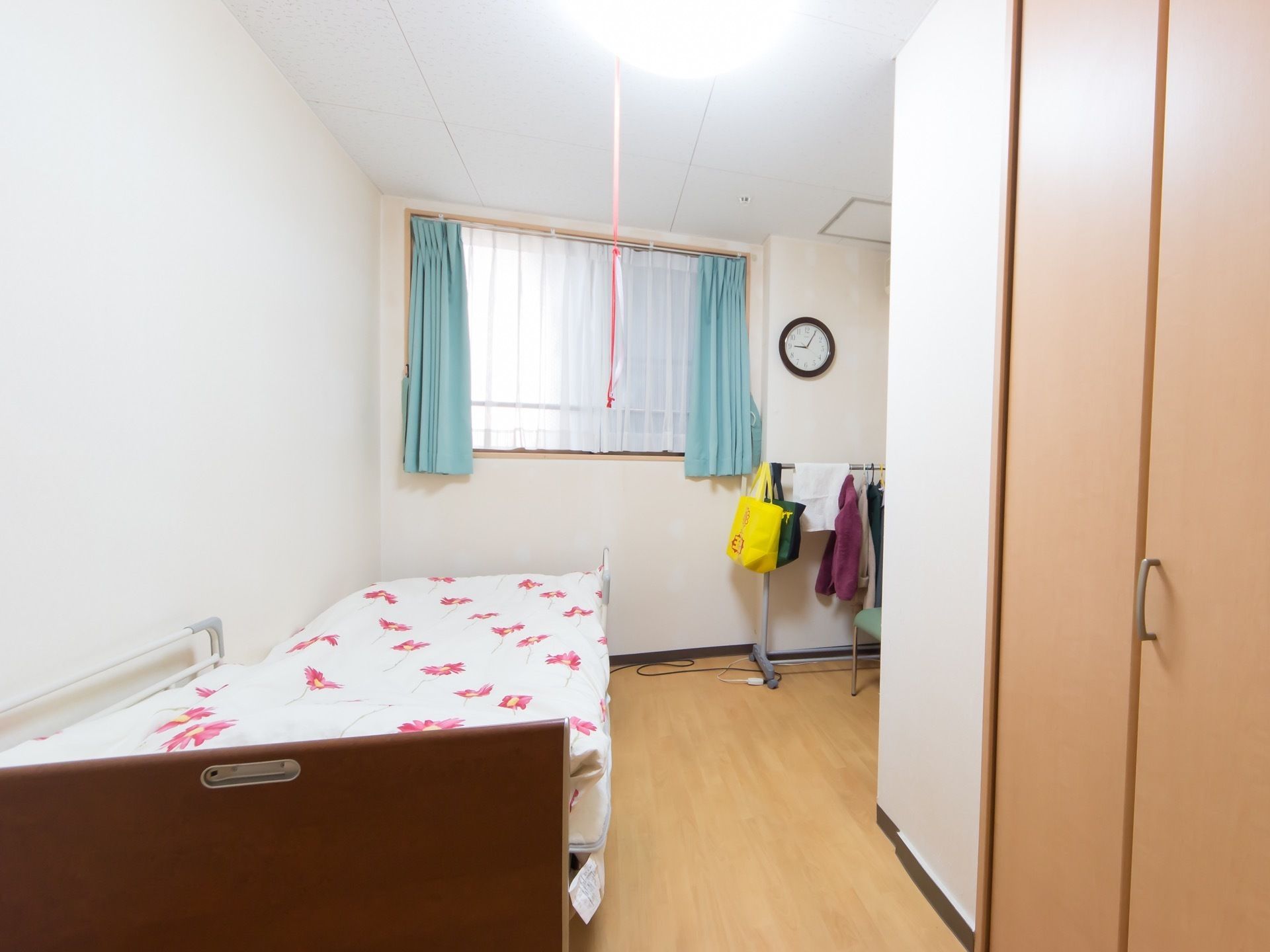個室タイプの居室である。左手前にベッドがある。奥にカーテンがかかった窓があり、その前にハンガーラックがある。