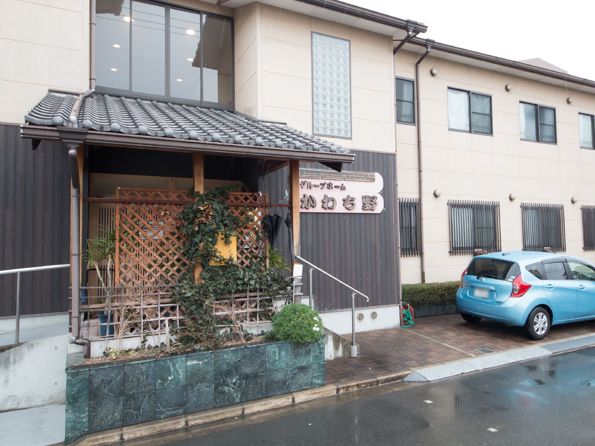 和風の装飾が施された玄関で、玄関ポーチに瓦屋根がせり出している。入口横に水色の乗用車が１台停まっている。