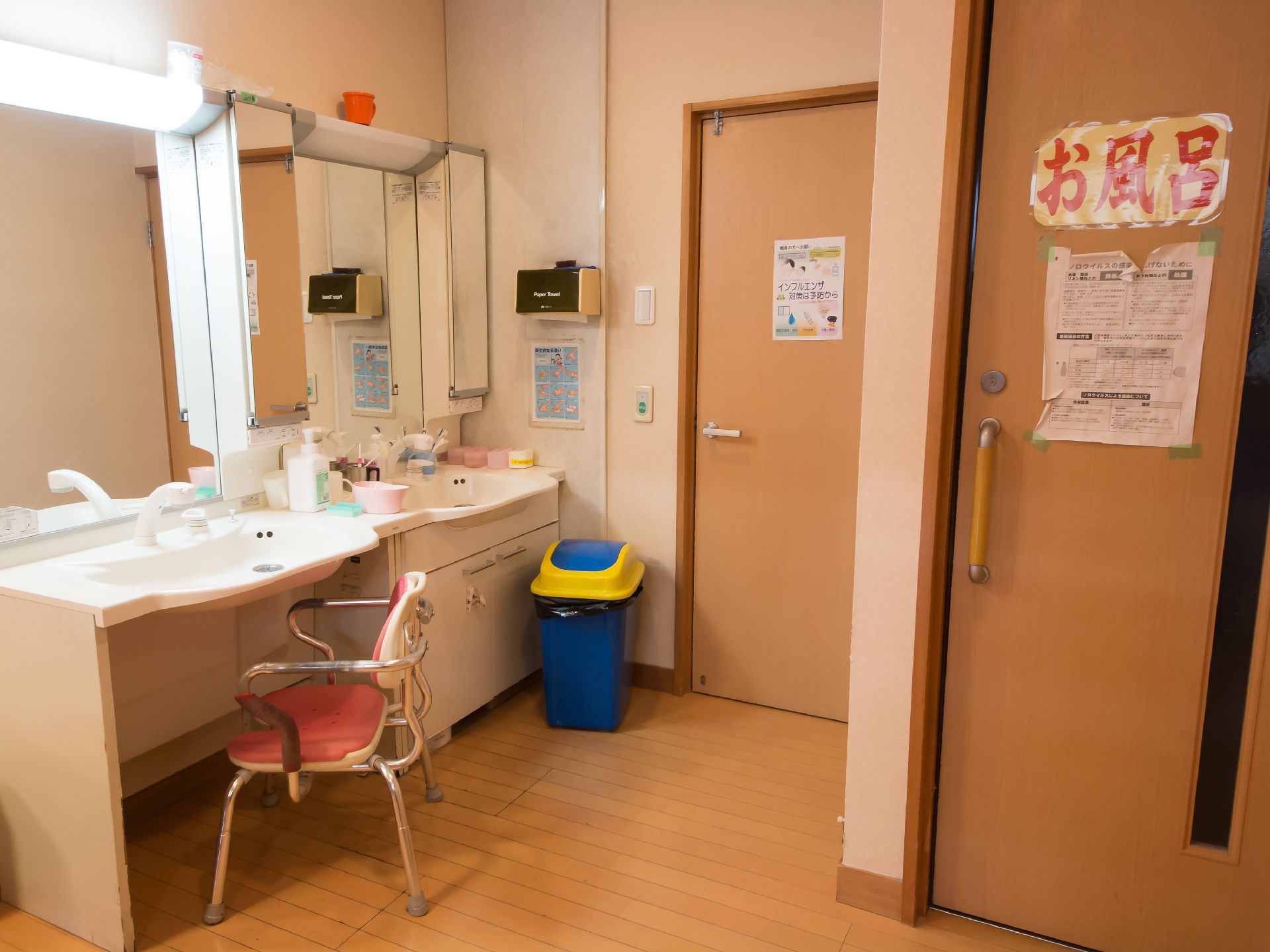 左奥に洗面台が２か所にあり、片方は鏡の上の照明が点灯している。流し前に椅子が置かれ、壁際にゴミ箱がある。