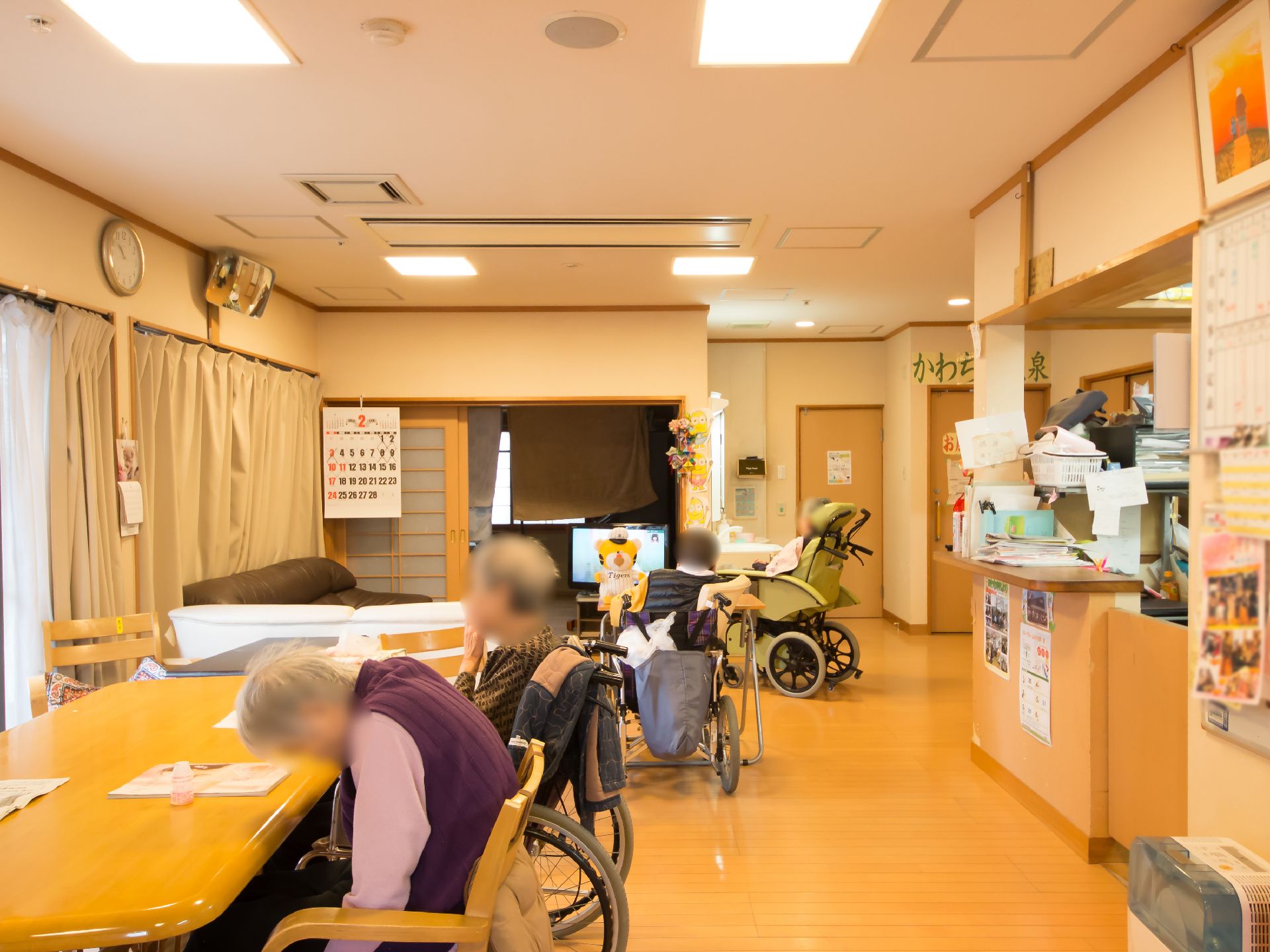 フローリングの床の共有スペースで、入居者様がダイニングテーブルでくつろいでいる。テレビ鑑賞をする入居者様もいる。