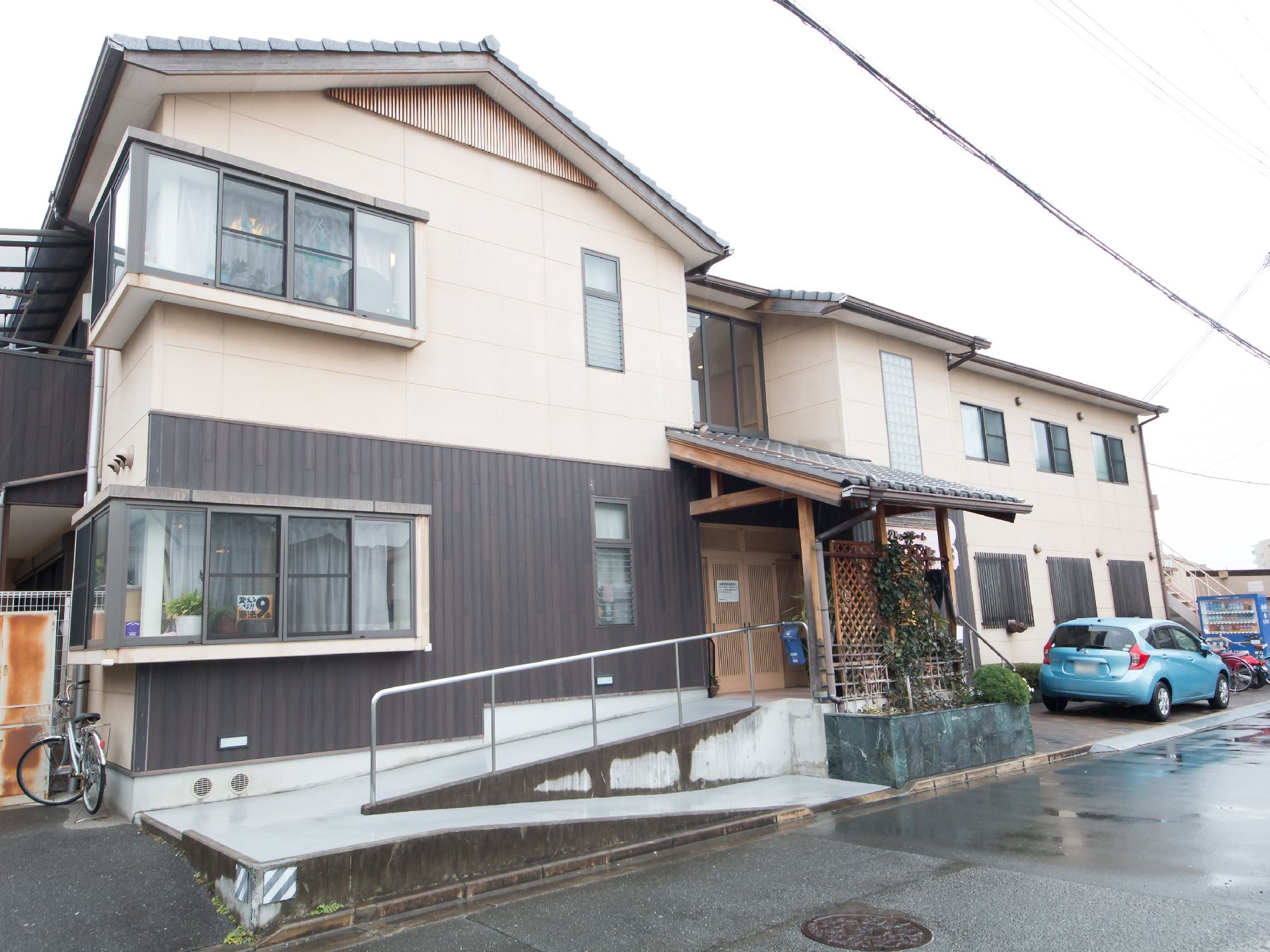 一般住宅のような三角屋根が乗った２階建ての建物である。施設前に自動車が１台停まっている。駐車場から玄関までスロープがある。