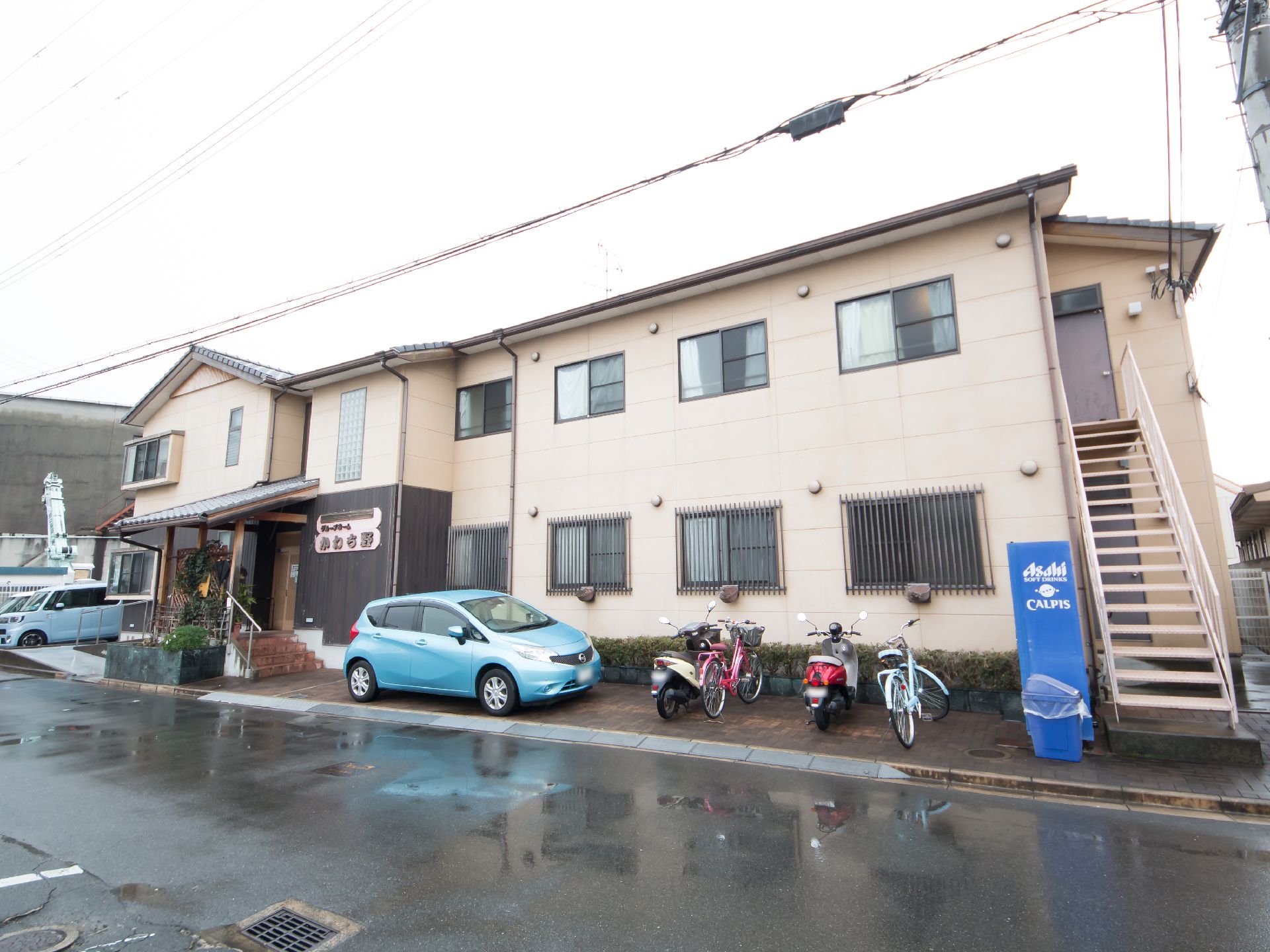 ベージュの外壁の建物で２階建てになっている。外階段があり、施設前には自転車が並び、自動販売機が置かれている。