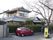 一部２階建ての施設である。植栽が敷地境界の垣根になっている。玄関横に、一台分の駐車スペースがあり、赤色の軽自動車が停まっている。