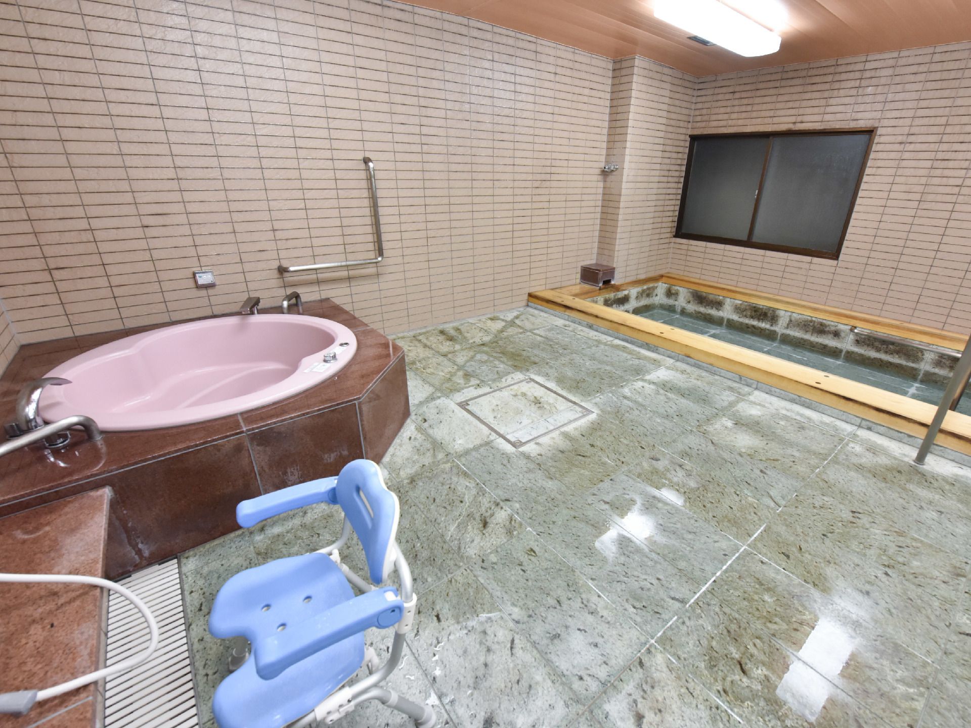 ピンクで円形タイプの浴槽はスイッチでジャグジーを楽しむことができる。壁には手すりも設置されている。手前にはシャワーがあり椅子も置かれている。
