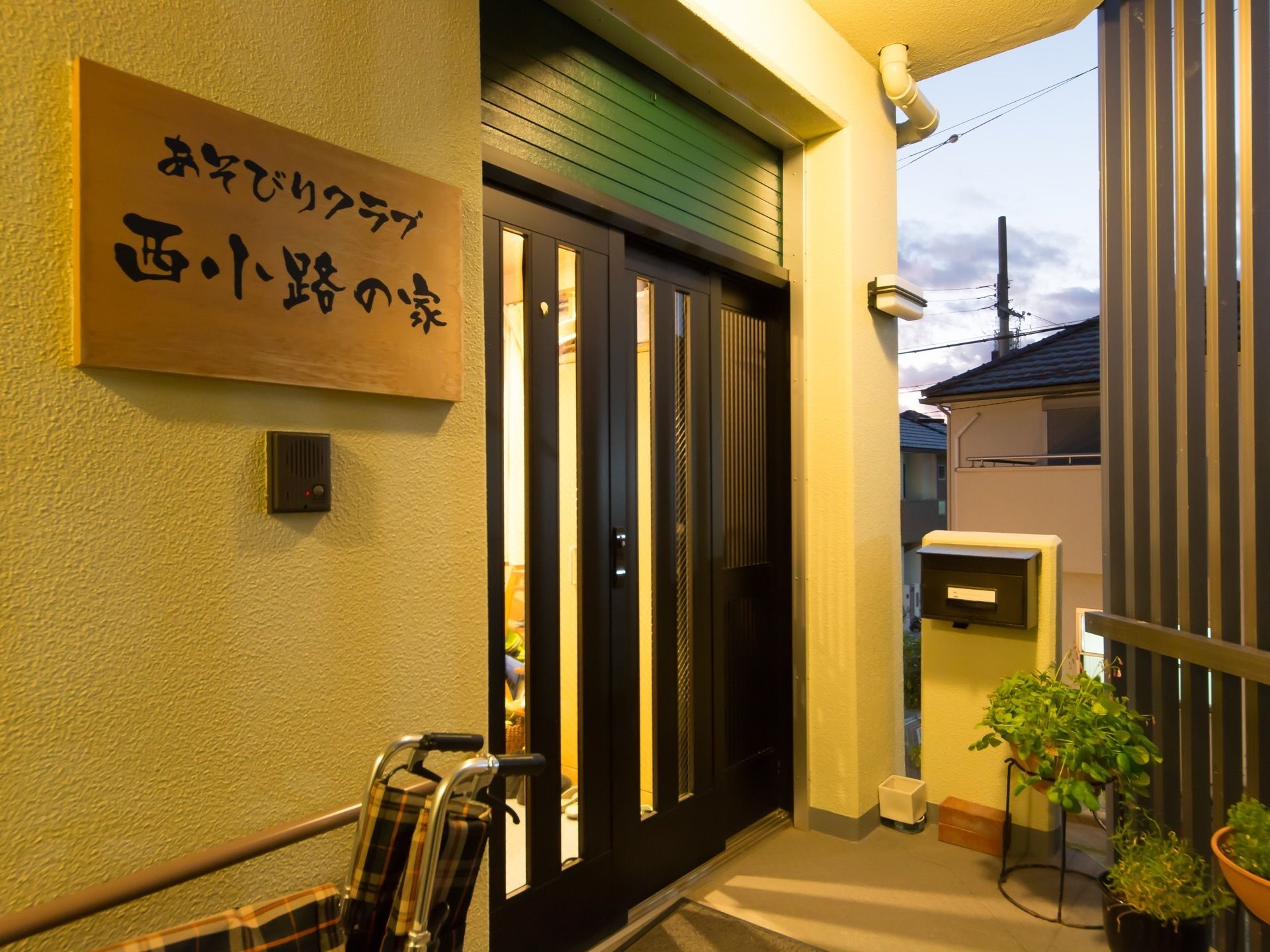 一般住宅のような玄関が設えられている。スライド式扉横の外壁には、木の板に施設名称が明記され設置されている。