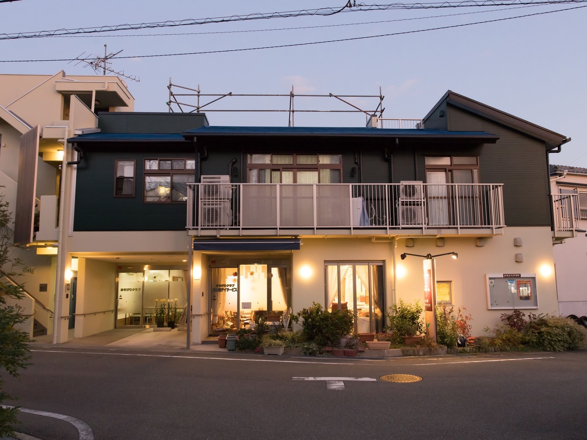 ベランダや屋上を完備した2階建て施設の外観である。夕暮れ時で照明器具が点けられ、温かみある印象である。