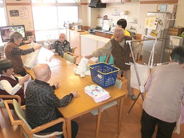ダイニングテーブルを囲んで洗濯物を畳む男女の入居者。窓が大きく明るい雰囲気。