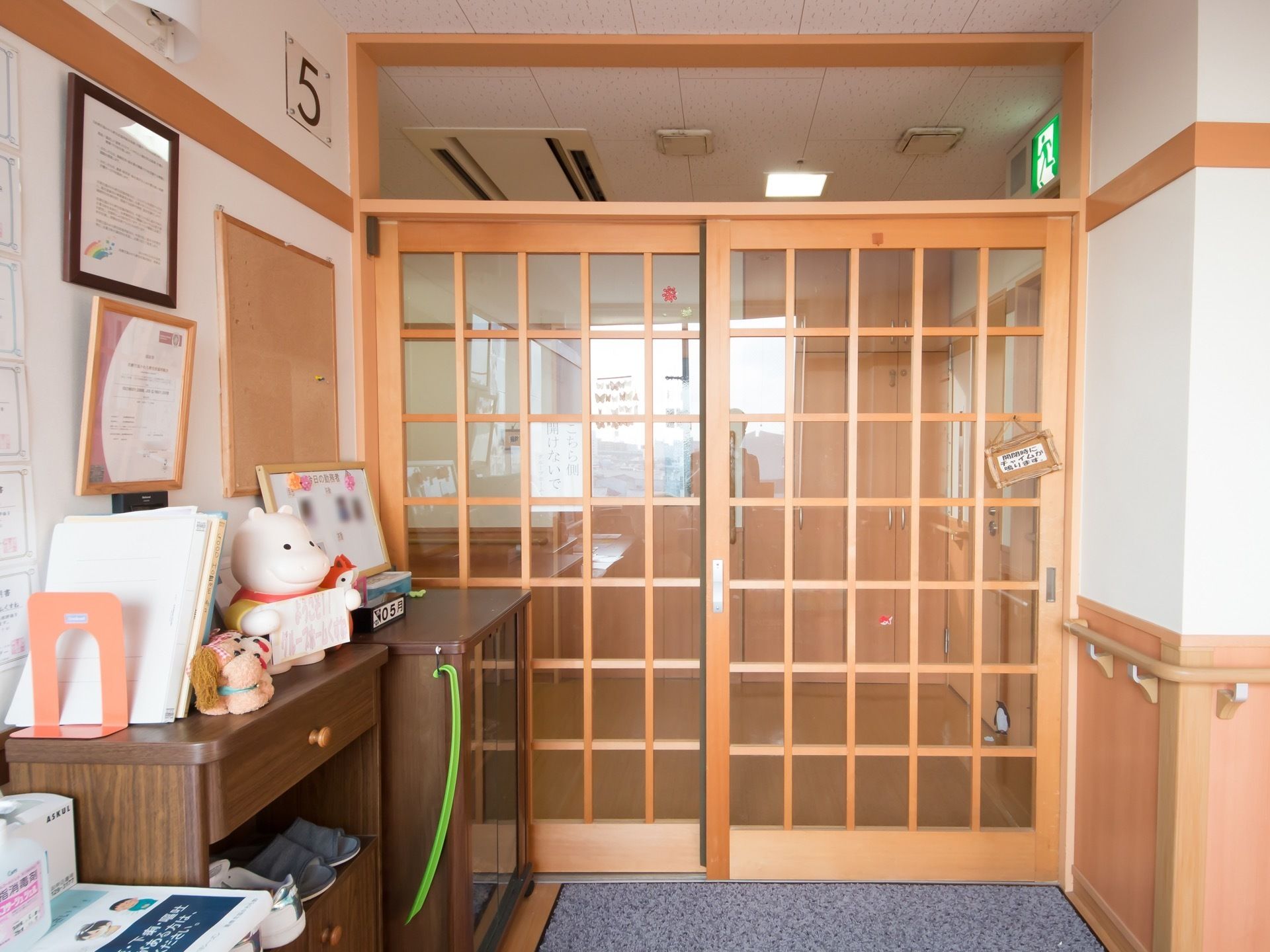 木の素材を使用した和風の横スライド式ドアになっている。ドアの前には下駄箱が設置されており、壁には掲示物が貼り出されている。