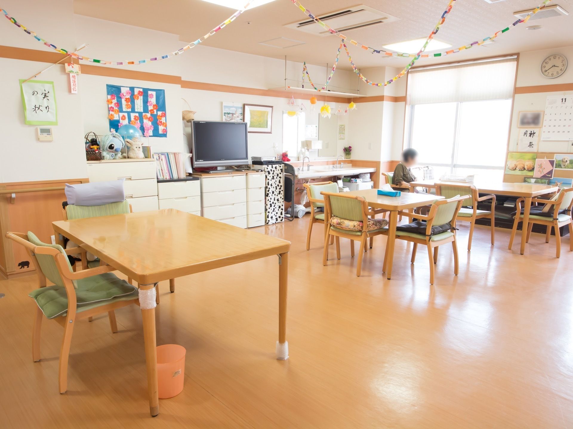 テレビや雑誌が用意されていて、一人の入居者様が椅子に腰かけて本を読んでいる。壁には手作りの装飾や絵画が飾られている。