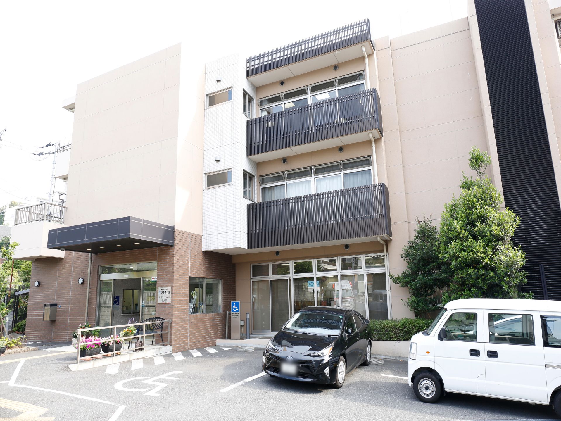 施設は白と茶色の3階建ての建物になっている。入り口の横は駐車場で車が3台停められる。車椅子用の駐車スペースも見える。