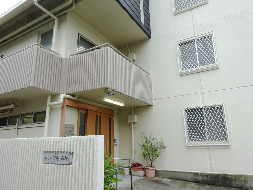 玄関先には草木が植えられている。白い外壁の４階建ての建物の前には門があり、ホーム名が書かれている。