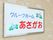 明るい色調の外壁に、施設名が書かれた看板が設置されている。背景色は白色、文字は青と赤色である。縁はグレー。