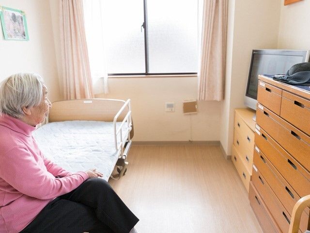 寝具付きのベッドの上には高齢女性が座っていて、タンスやテレビなどが置かれた部屋には大きな窓がある。