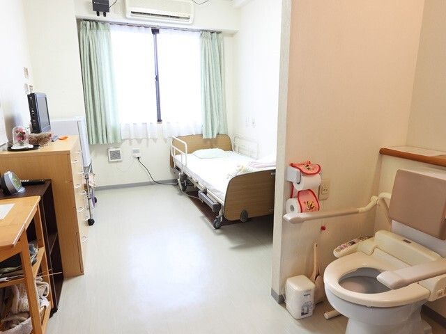 ドアのないトイレには手すりと背もたれが付いた便座が設置され、その奥にはベッドなどが置かれている。