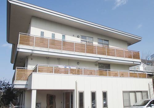 青い空の下3階建ての建物が堂々と建っている。白を基調にした建物に木製のベランダがアクセントになっている。