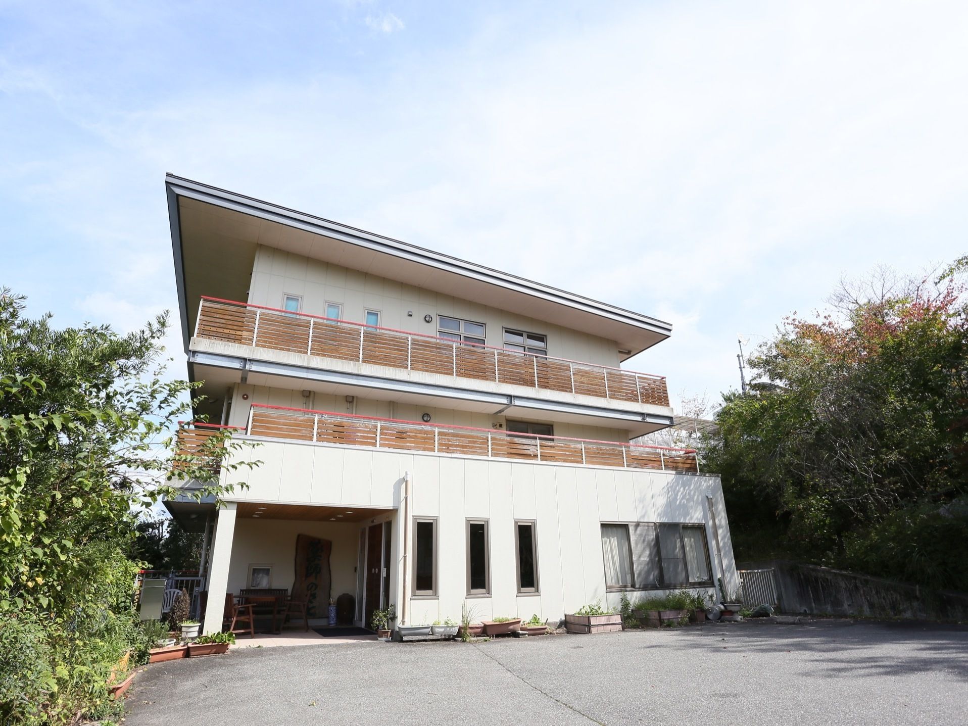 屋根付きの玄関先には、ベンチが置かれている。広い駐車場で、周りには草木が茂っている。白で統一された建造物である。