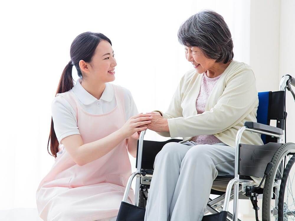 黄色いカーディガンを着た高齢女性が車いすに座り、そばにはエプロンを付けた若い女性スタッフがいて、顔を見合わせてほほ笑んでいる。